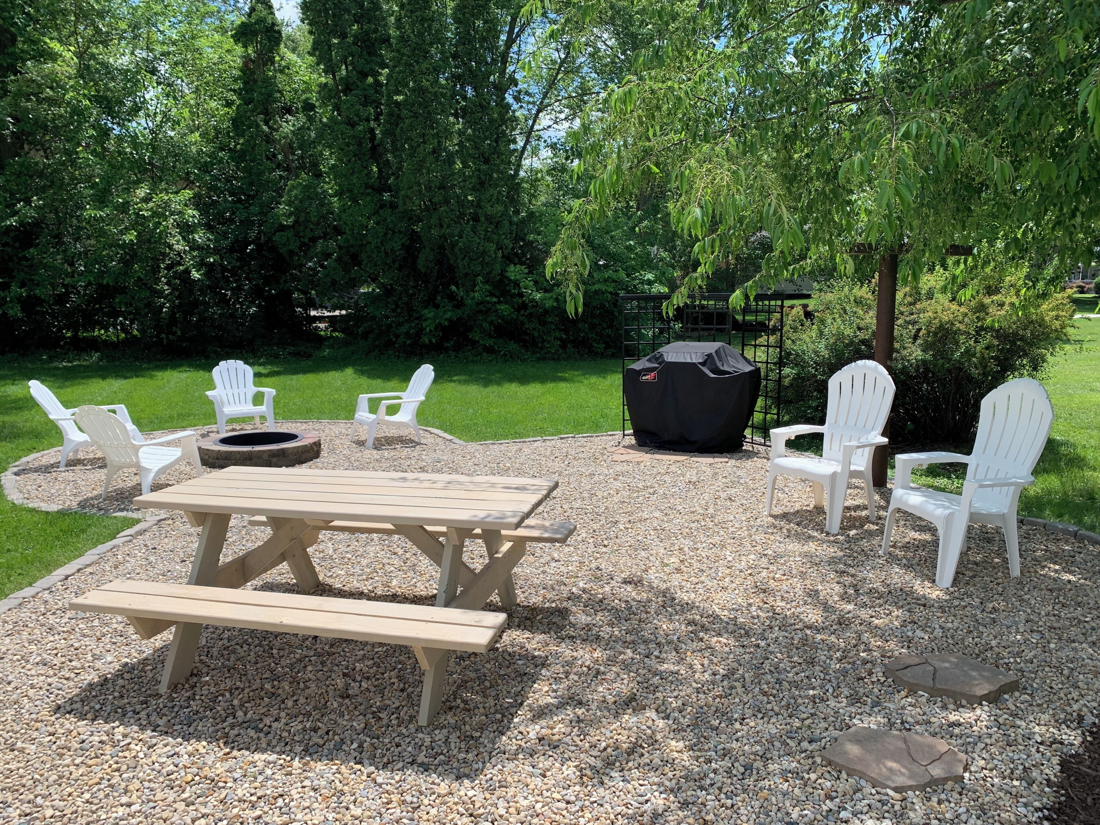 Een terras buiten met een picknicktafel, witte Adirondack stoelen en een barbecue.