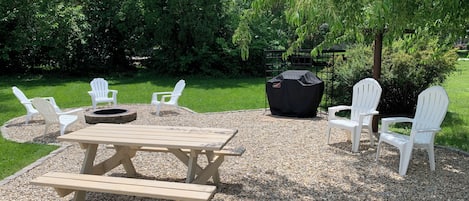 Een terras buiten met een picknicktafel, witte Adirondack stoelen en een barbecue.
