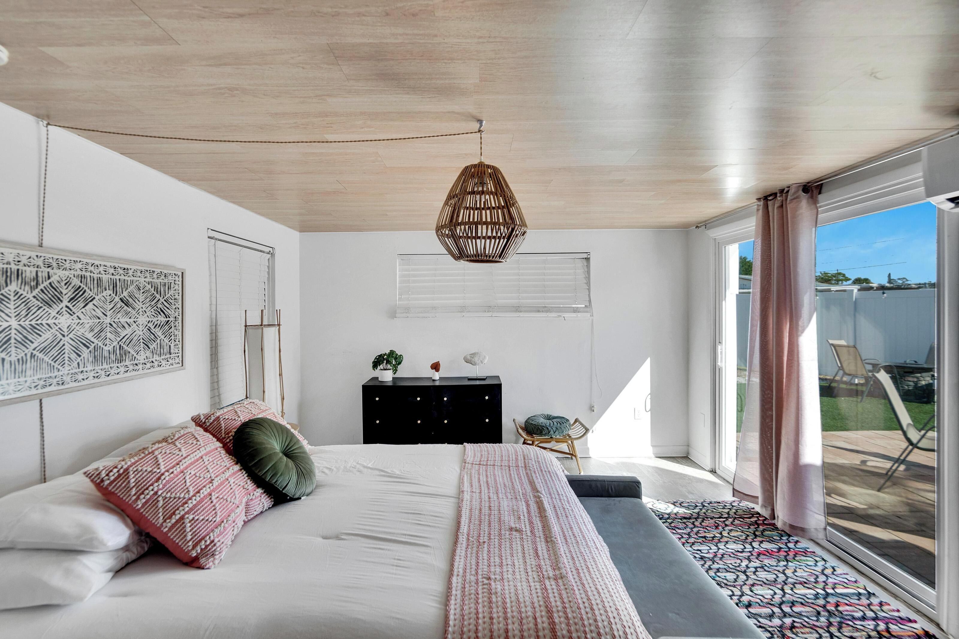 A bedroom with a bed, a dresser, a hanging light fixture, and a view through a window.