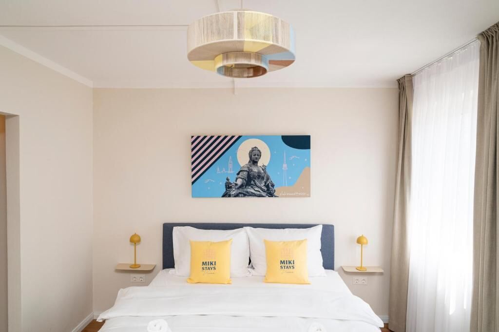 Ein Hotelzimmer mit einem Bett, zwei Nachttischlampen, einem Wandbild und einem Fenster mit Vorhängen.