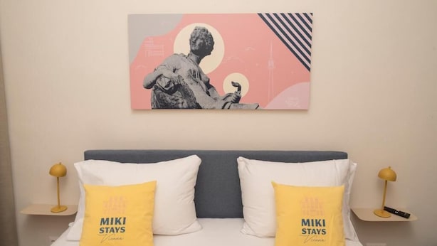 A hotel room with a bed, two pillows with 'MIKI STAYS Vienna' text, a wall-mounted artwork, and bedside tables with lamps.