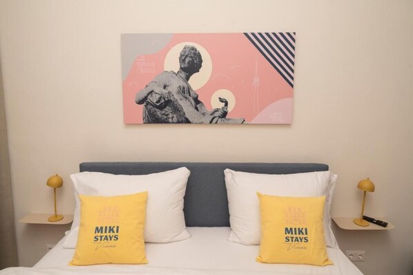 A hotel room with a bed, two pillows with 'MIKI STAYS Vienna' text, a wall-mounted artwork, and bedside tables with lamps.