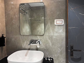 A bathroom with a white sink, a mirror, and a wall-mounted light switch. - Shivlok Palace (Varanasi)