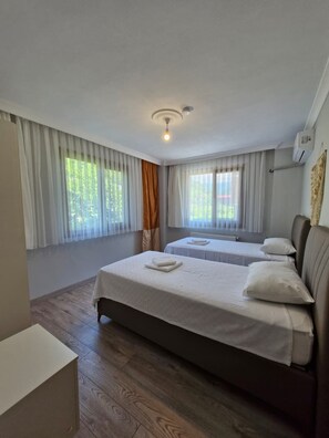 A hotel room with two beds, a wooden floor, a ceiling light, and a window with curtains.