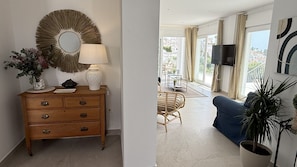 A living room with a wooden dresser, a mirror, a lamp, a blue sofa, a television, and a view of the outdoors through large windows. - Nerja Paradise Rentals - Villa Barranco (Nerja)