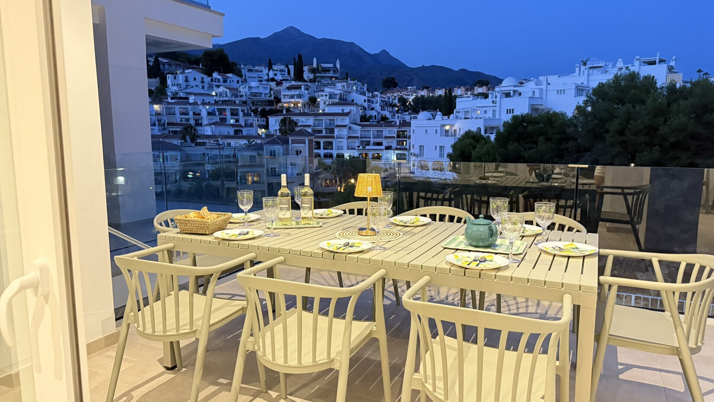 Mesa lista para cuatro personas, con sillas, vista a un pueblo en una ladera y una montaña al fondo.
