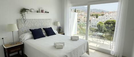 A hotel room with a large bed, two bedside tables, a nightstand, a window with a view of a cityscape, and a balcony with chairs.