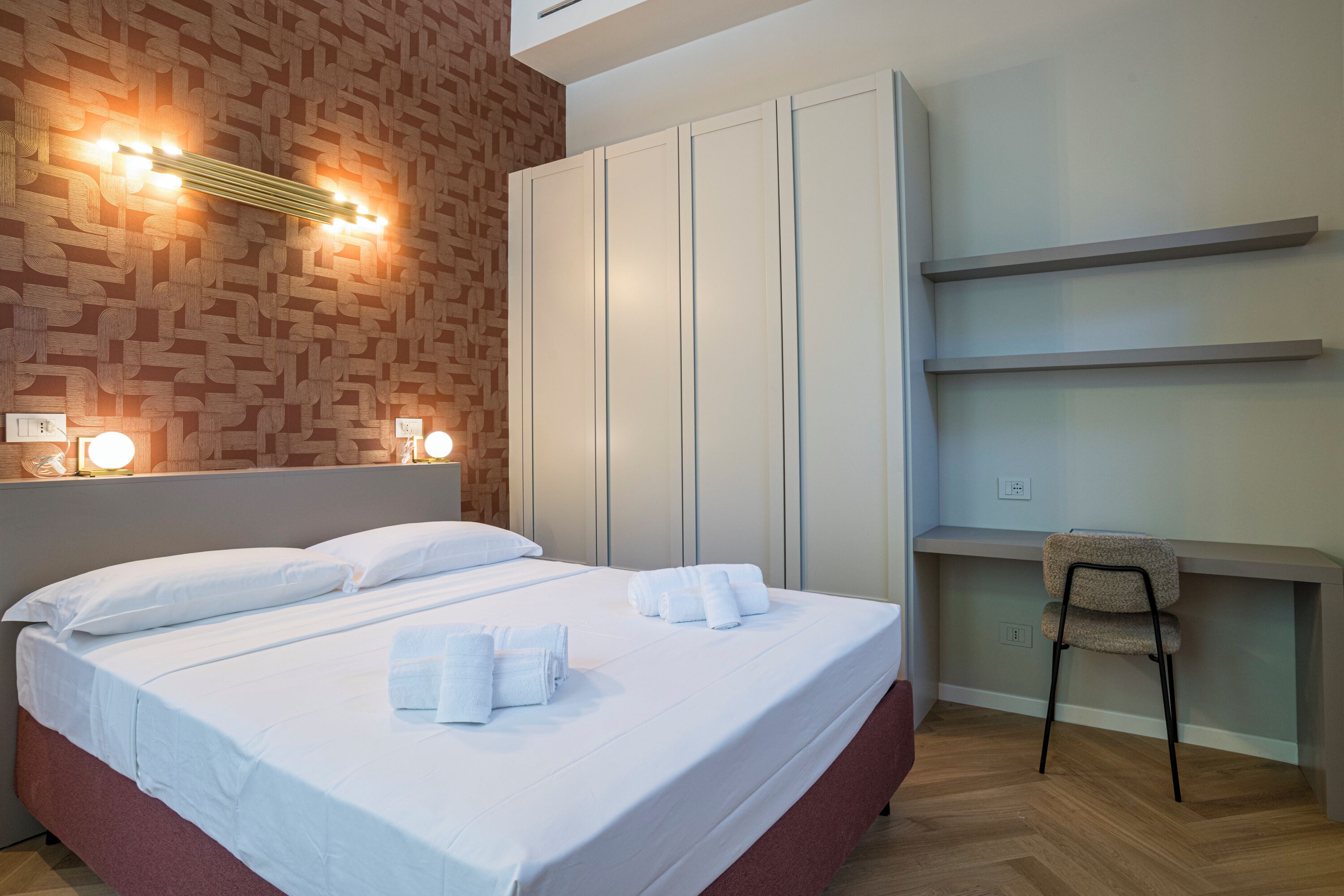 Una habitación de hotel con cama, lámparas de noche, armario, escritorio y una silla.