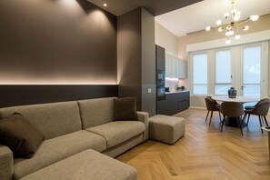 A modern living room with a sectional sofa, a dining area with a round table and chairs, and a kitchen with dark cabinetry.