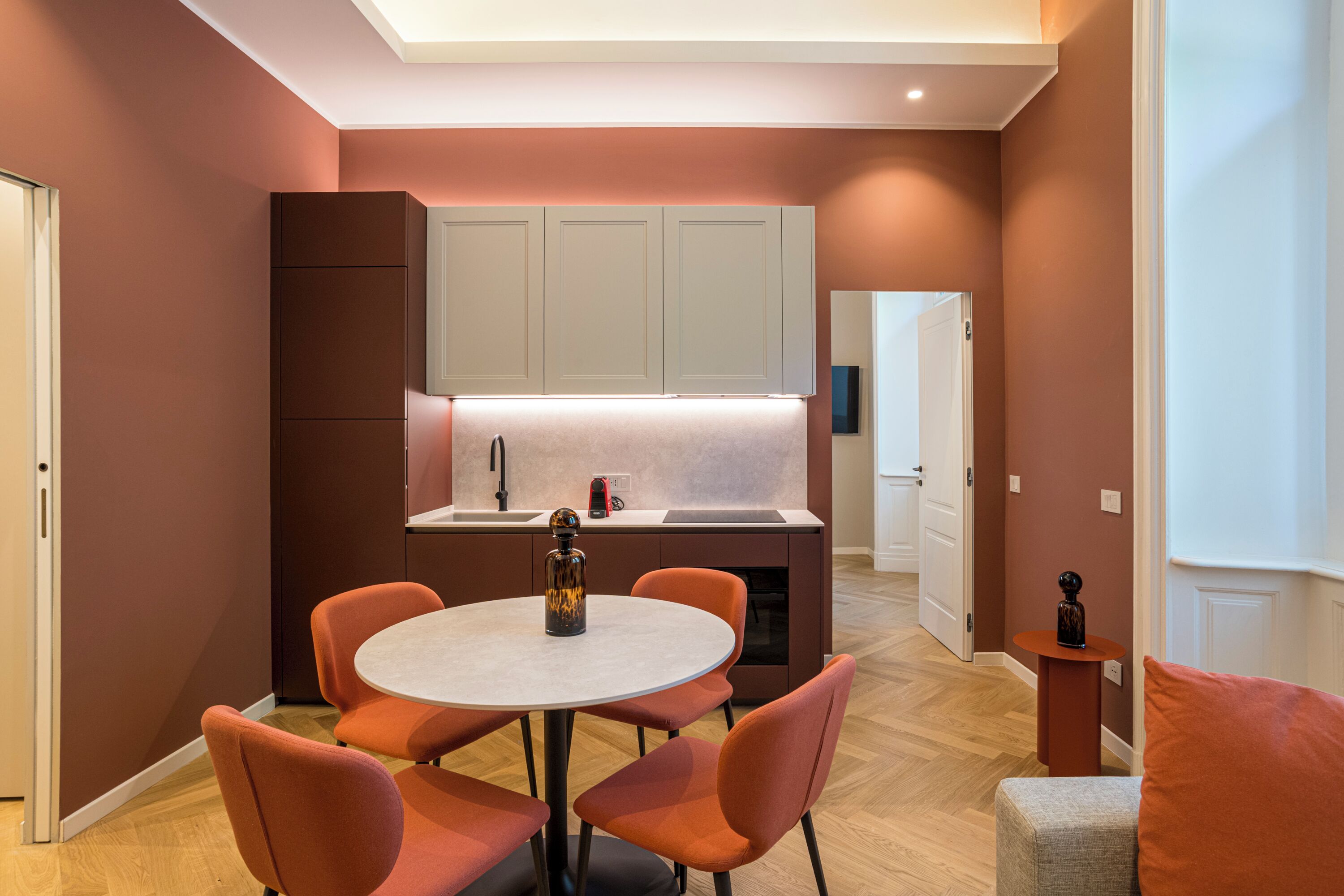 A compact kitchen with a dining area, featuring a round table and matching chairs, a built-in oven, and a countertop with a sink and a red appliance.