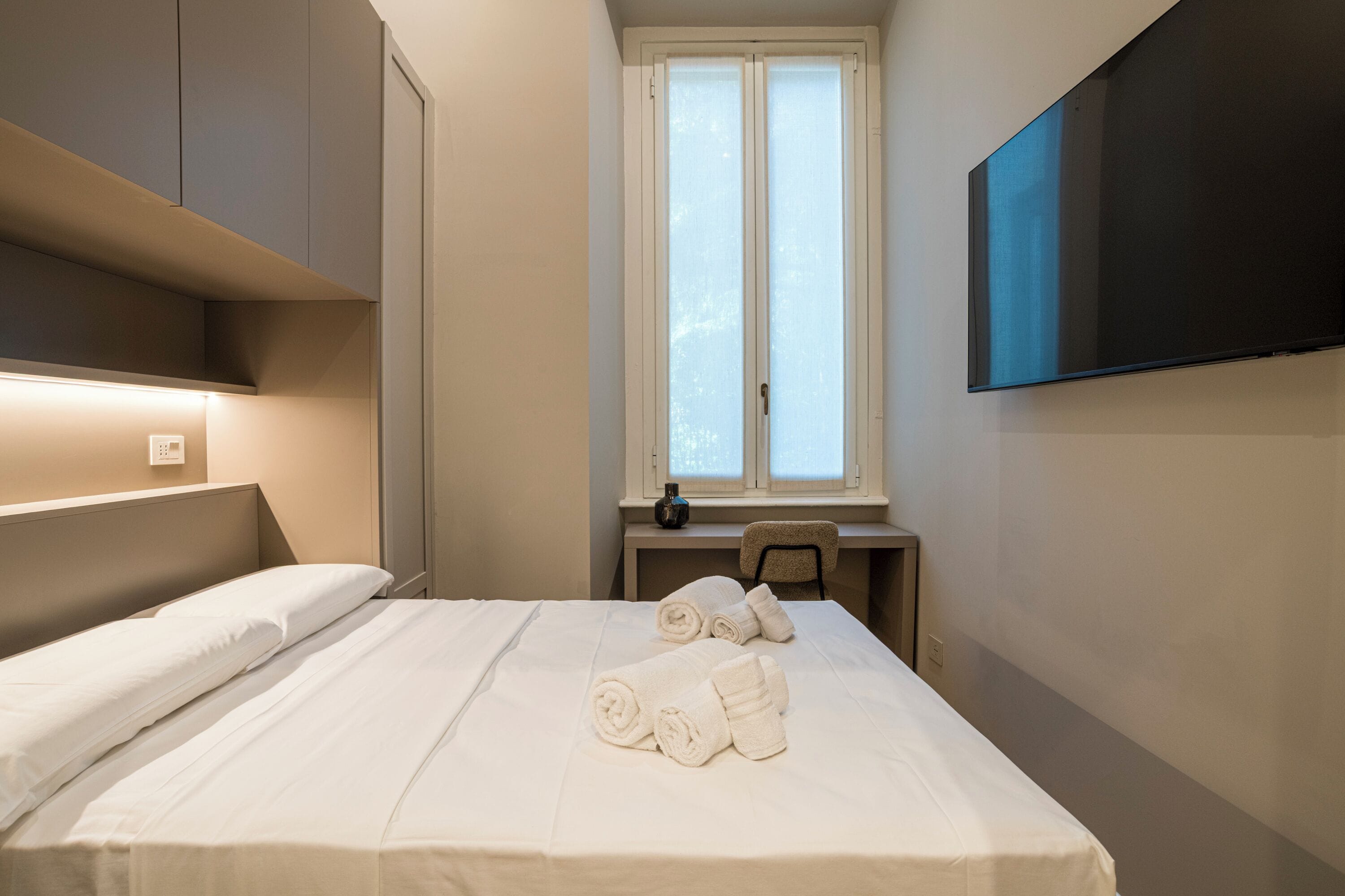 A hotel room with a bed, a desk, a chair, a window, and a TV mounted on the wall.