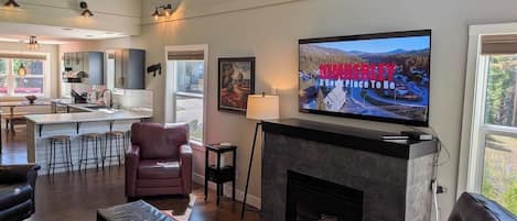 Ein modernes Wohnzimmer mit Kamin, einem darüber montierten Fernseher und einer Küche im Hintergrund.