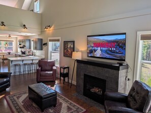 A modern living room with a fireplace, a television mounted above it, and a kitchen in the background. - Spacious Kimberley Mountain Retreat | 5 Bedrooms (Kimberley)