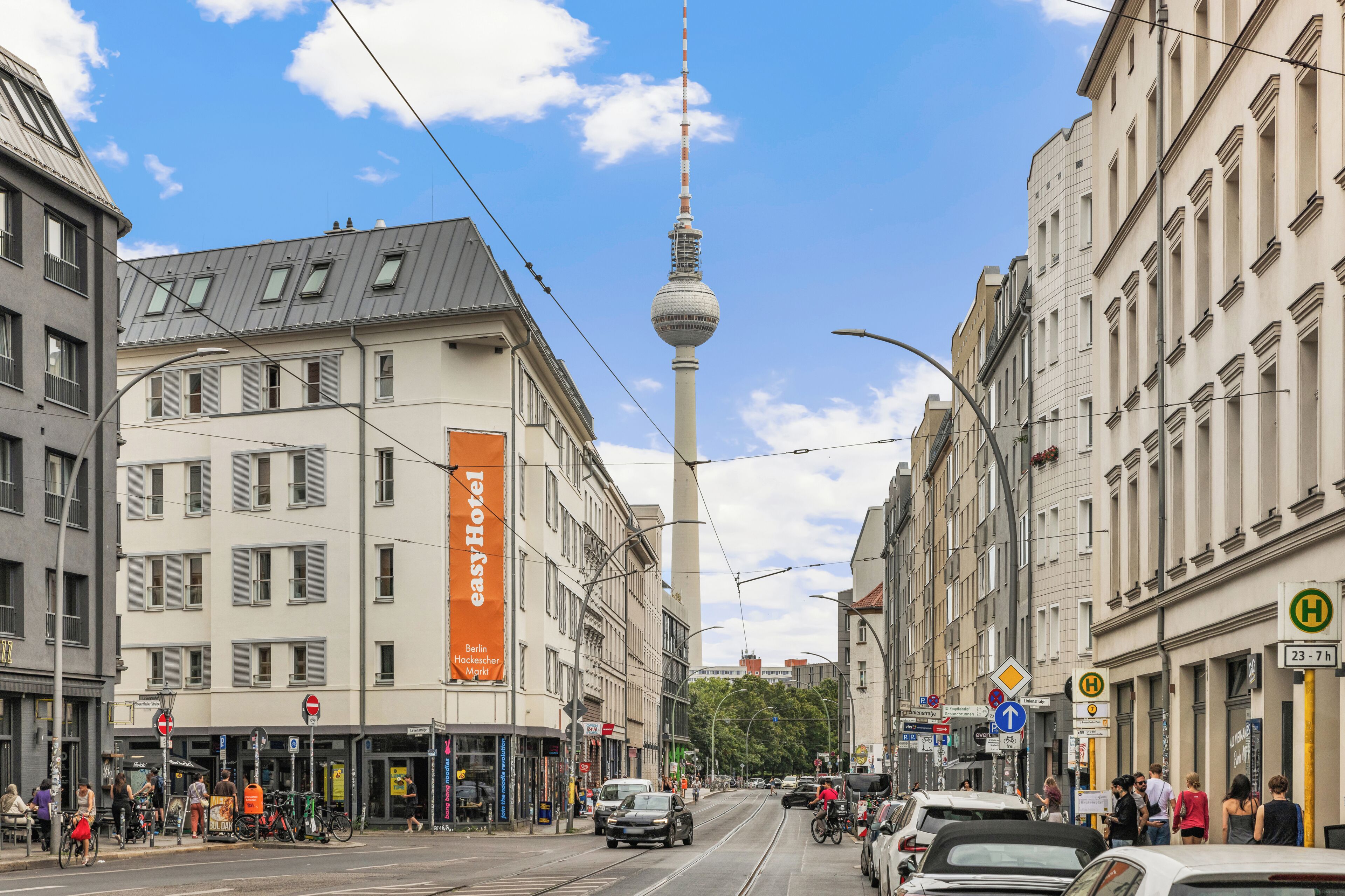 Straßenansicht mit einem markanten Turm im Hintergrund, einem Gebäude mit einer großen orangefarbenen Werbetafel und verschiedenen Geschäften sowie Fußgängern.