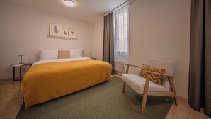 A bedroom with a bed, a chair, a nightstand, a window with curtains, and wall-mounted artwork.