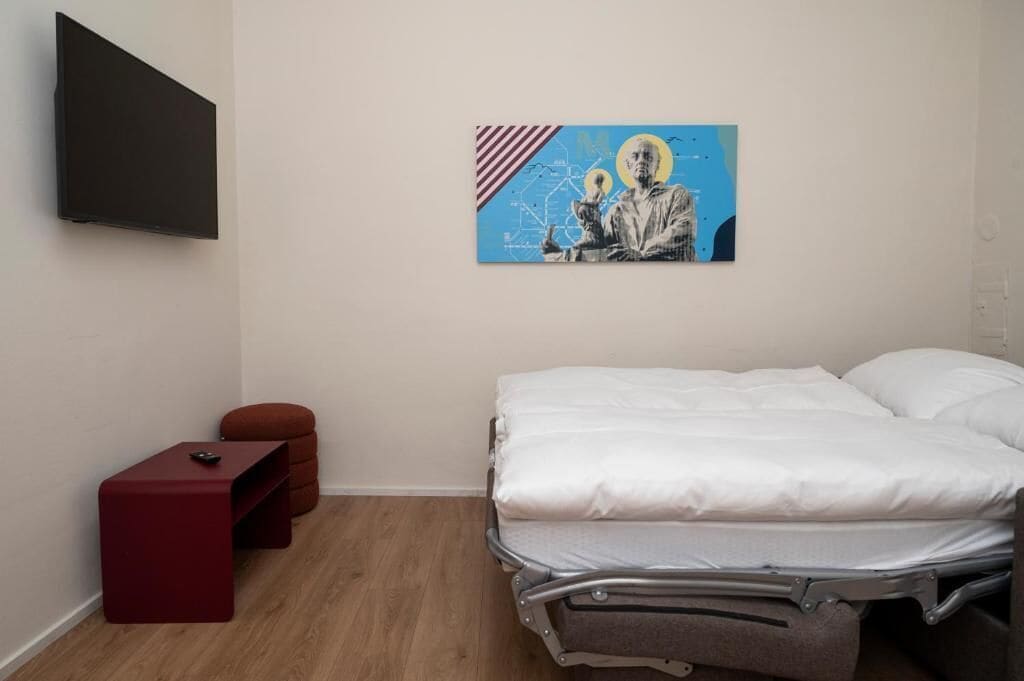 Un dormitorio con una cama, un reposapiés rojo, una mesita y un televisor montado en la pared.