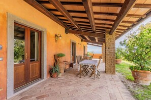Eine Terrasse im Freien mit einer Holz-Pergola, einem Tisch mit Stühlen, Blumentöpfen und Blick auf grüne Vegetation.