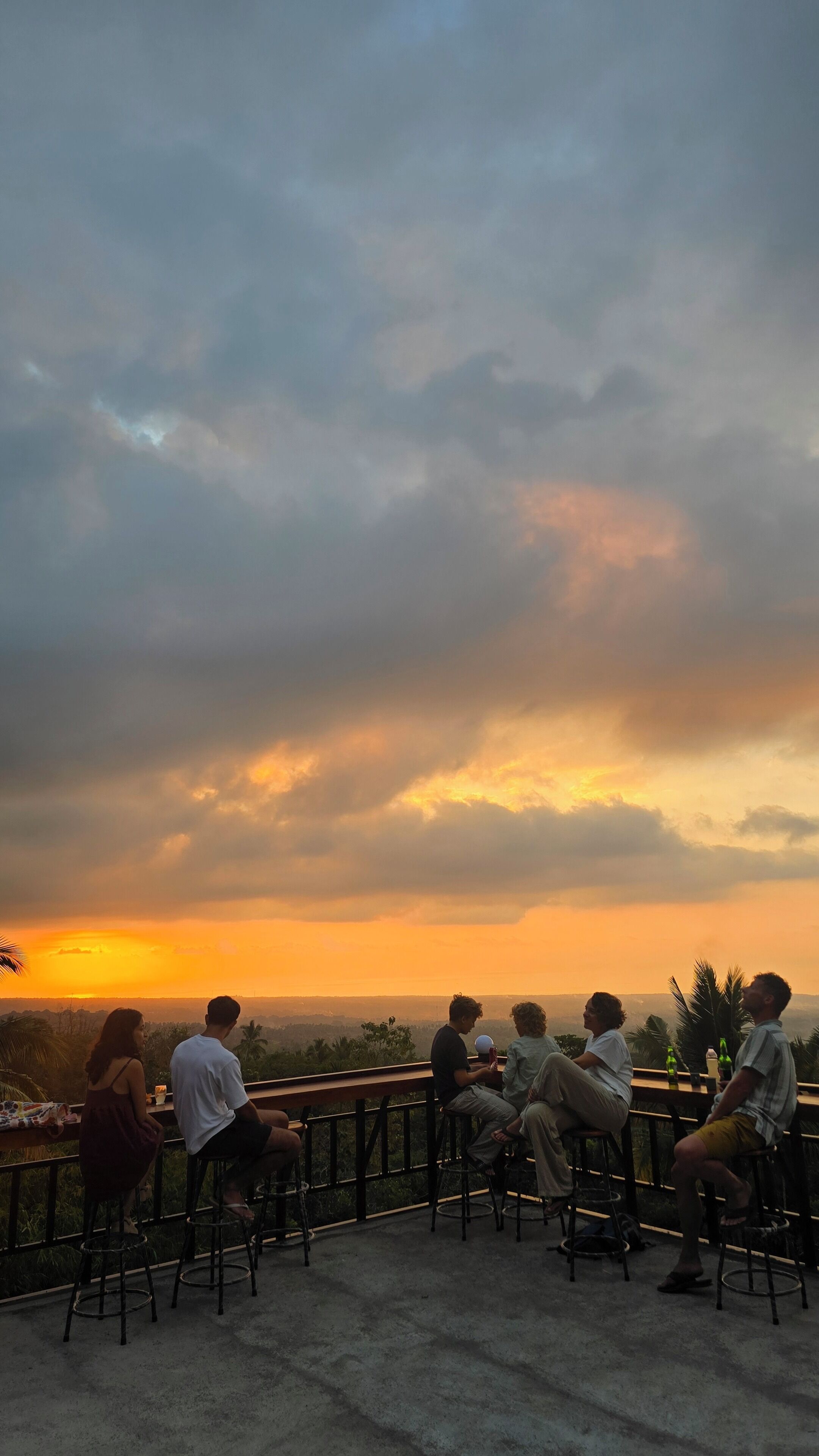 A group of people seated on outdoor chairs with a view of the sunset over a landscape.