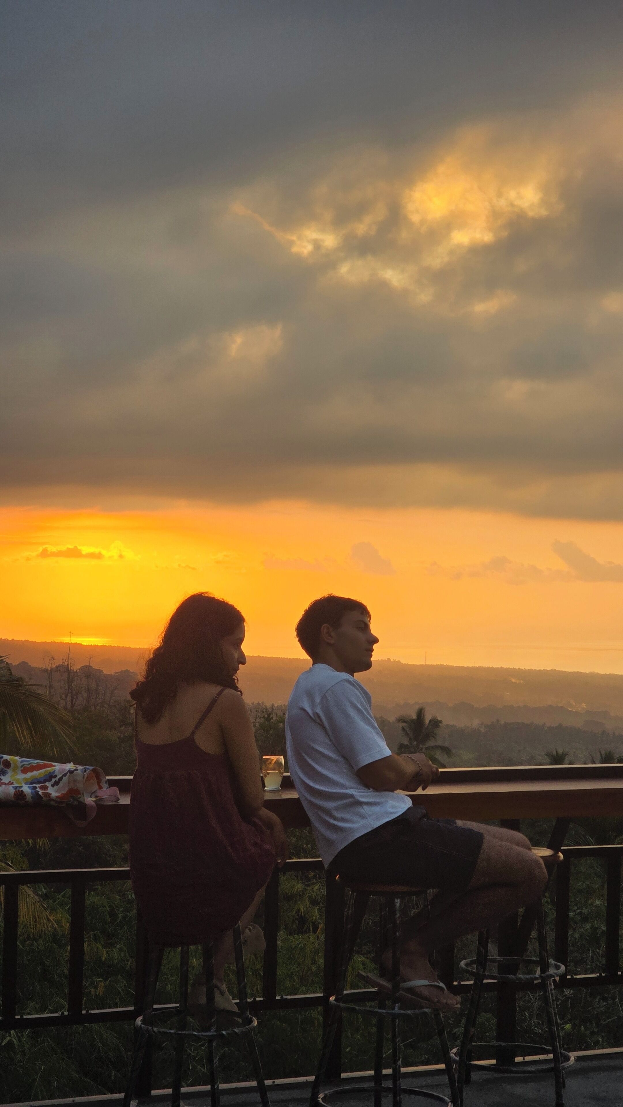 Twee personen zitten op een balkon met uitzicht op een zonsondergang en het landschap.