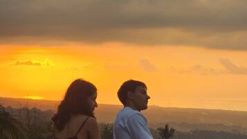 Twee personen zitten op een balkon met uitzicht op een zonsondergang en het landschap.
