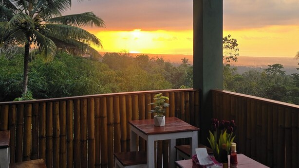 An outdoor seating area with tables and chairs, overlooking a landscape with palm trees and a sunset.