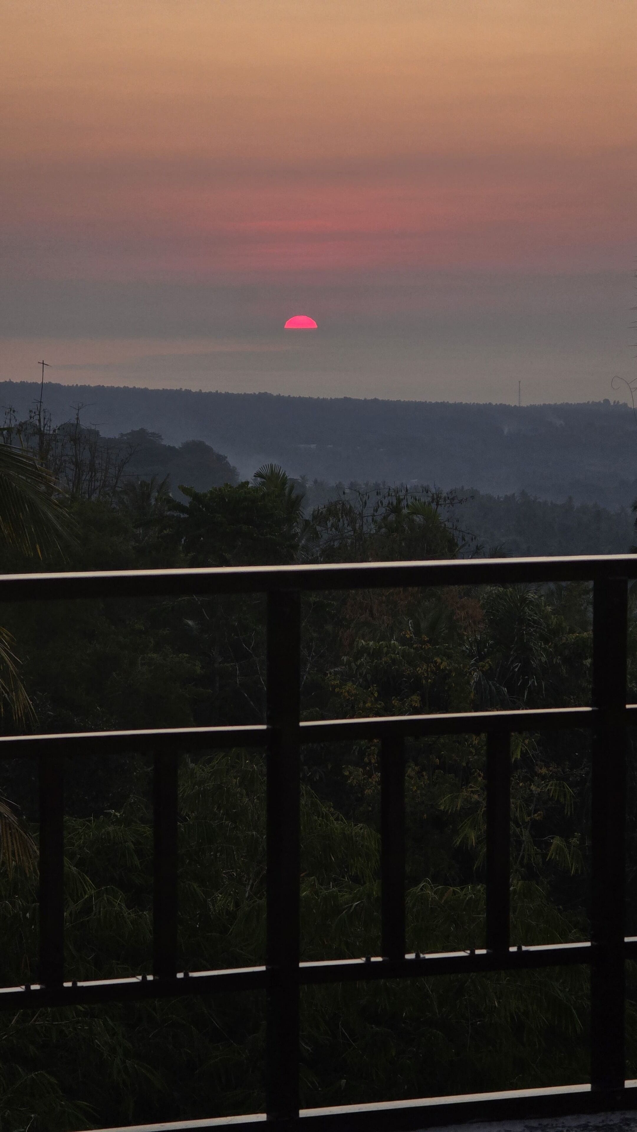 A sunset view from a balcony with a railing, overlooking a forested landscape.