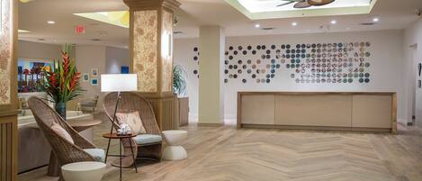 Un hall d’entrée doté d’un parquet en bois à motifs chevrons, de meubles en osier, d’un comptoir de réception et d’un mur recouvert de miroirs ronds.