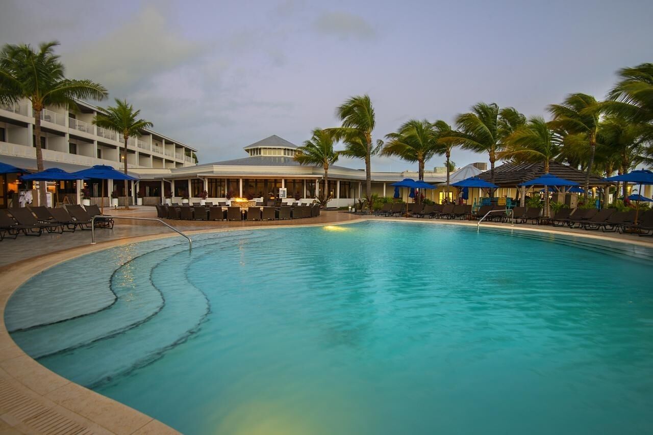 A swimming pool with lounge chairs and umbrellas, surrounded by palm trees and a hotel building.