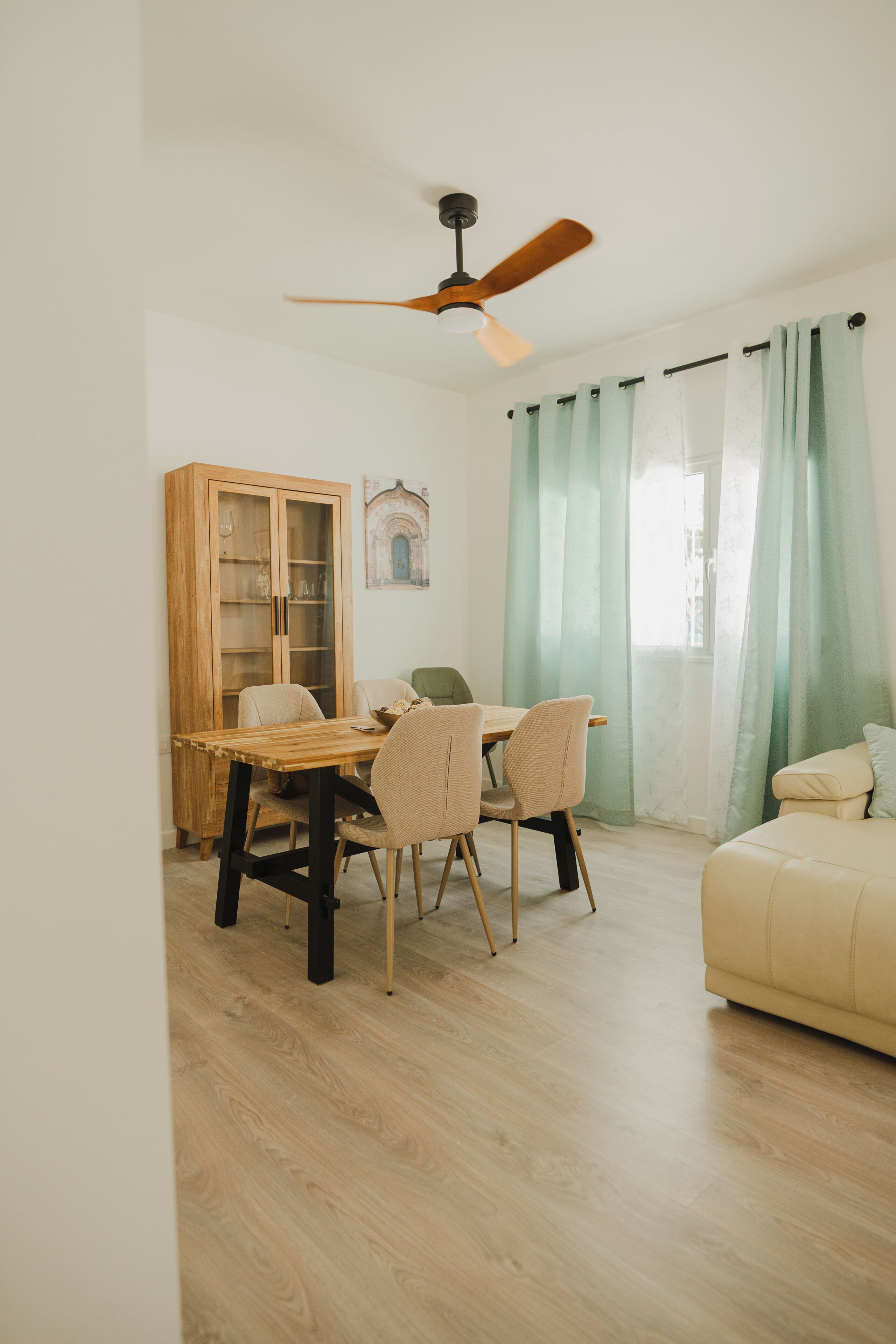 A room with a wooden dining table, chairs, a cabinet with glass doors, a ceiling fan, and a sofa.
