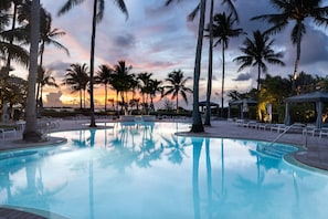 Ein Pool mit klarem Wasser, umgeben von Palmen und Liegestühlen, vor einem Sonnenuntergangshimmel.