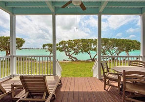 Ein überdachter Außenbereich mit Holzdeck, weißen Geländern und Blick auf das Meer und Bäume.