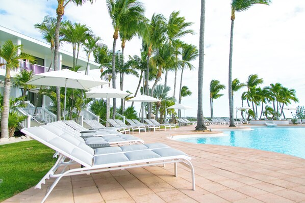 A poolside area with lounge chairs and umbrellas, surrounded by palm trees and a building with balconies. - A Place You'll Surely Enjoy! Full Kitchen, Outdoor Swimming Pool Near Key West (Duck Key)