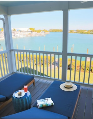 A covered outdoor seating area with blue cushions, a small round table with a drink and a hat, and a view of a calm body of water and distant buildings. - Unwind with Pool Access and Kitchen Near Lagoon | Duck Key Escape (Duck Key)