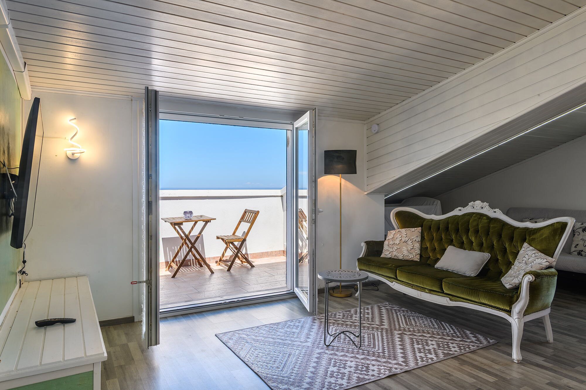 A room with a green sofa, a patterned rug, and a balcony with a table and chairs.
