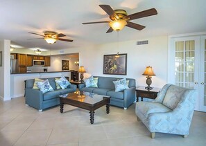 A living room with two sofas, a coffee table, and a patterned armchair. There are ceiling fans and two lamps. A kitchen is visible in the background. - Fully Equipped Kitchen and Pool Access Make This an Easy Choice for Your Stay (Duck Key)