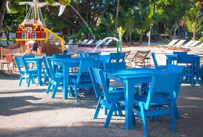 Outdoor seating with blue chairs and tables, a small boat structure, and a sandy area with lounge chairs in the background. - Charming Villa with Pool Access & Full Kitchen for Easy Meals! (Duck Key)