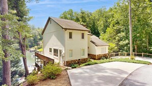 A two-story house with a sloped roof, a front porch, and a driveway leading to a dock. - Lake Lure Lakefront House, Private Dock, Multiple Decks, Kayaks, Mountain Views, Gameroom (Lake Lure)
