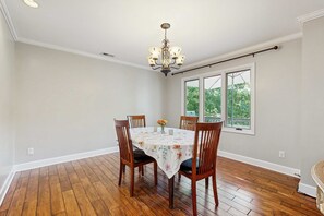 A dining room with a table set for four, wooden chairs, a chandelier, and a window with a view of greenery. - Lake Lure Lakefront House, Private Dock, Multiple Decks, Kayaks, Mountain Views, Gameroom (Lake Lure)