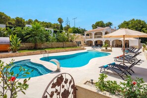 A swimming pool with lounge chairs and umbrellas, surrounded by greenery and a building with arches. - Villa El Silencio by Villa Plus (Jávea)
