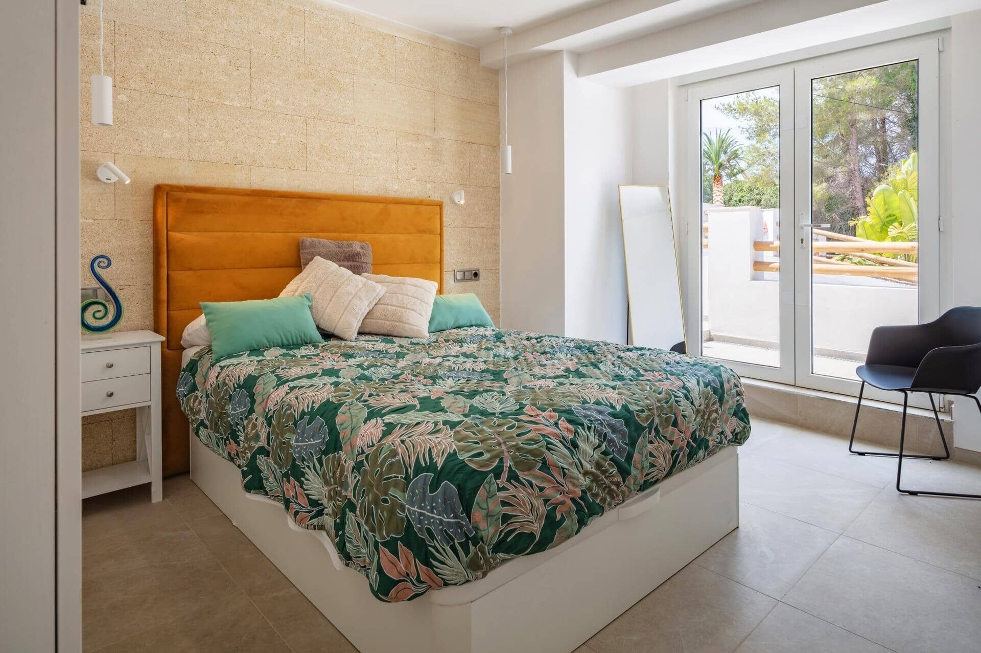 A bedroom with a bed, a nightstand, a chair, and a view of the outdoors through a glass door.