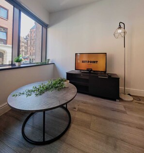 A modern living room with a round table, a television displaying 'HIT POINT Thursday 9pm', and a floor lamp. - City Centre One Bedroom Apartments (Liverpool)
