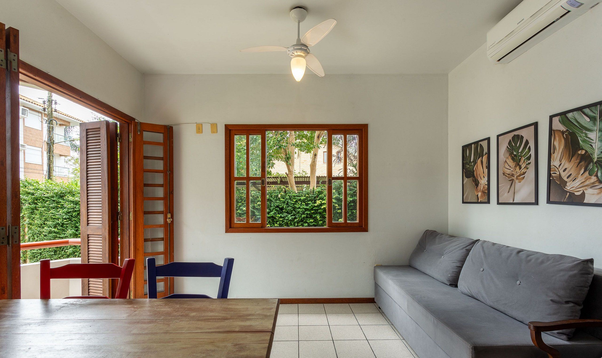 A room with a ceiling fan, a window with a view of greenery, a grey sofa, and a wooden table with chairs.