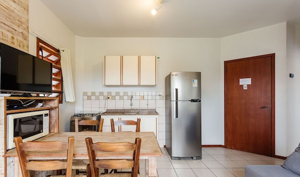 A compact kitchen with a TV, microwave, stove, and refrigerator. There is a dining table with chairs and a wooden door. - Charming Apt 270m From the Beach Stcy102 (Florianópolis)