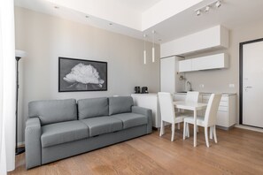 Een moderne woonruimte met een grijze bank, een witte eettafel met stoelen, een kitchenette en een gekaderd schilderij aan de muur.