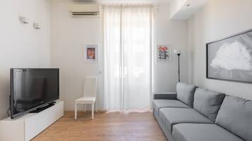 Een moderne woonkamer met een flatscreen televisie, een grijze bank, een witte stoel en een groot raam met dunne gordijnen.