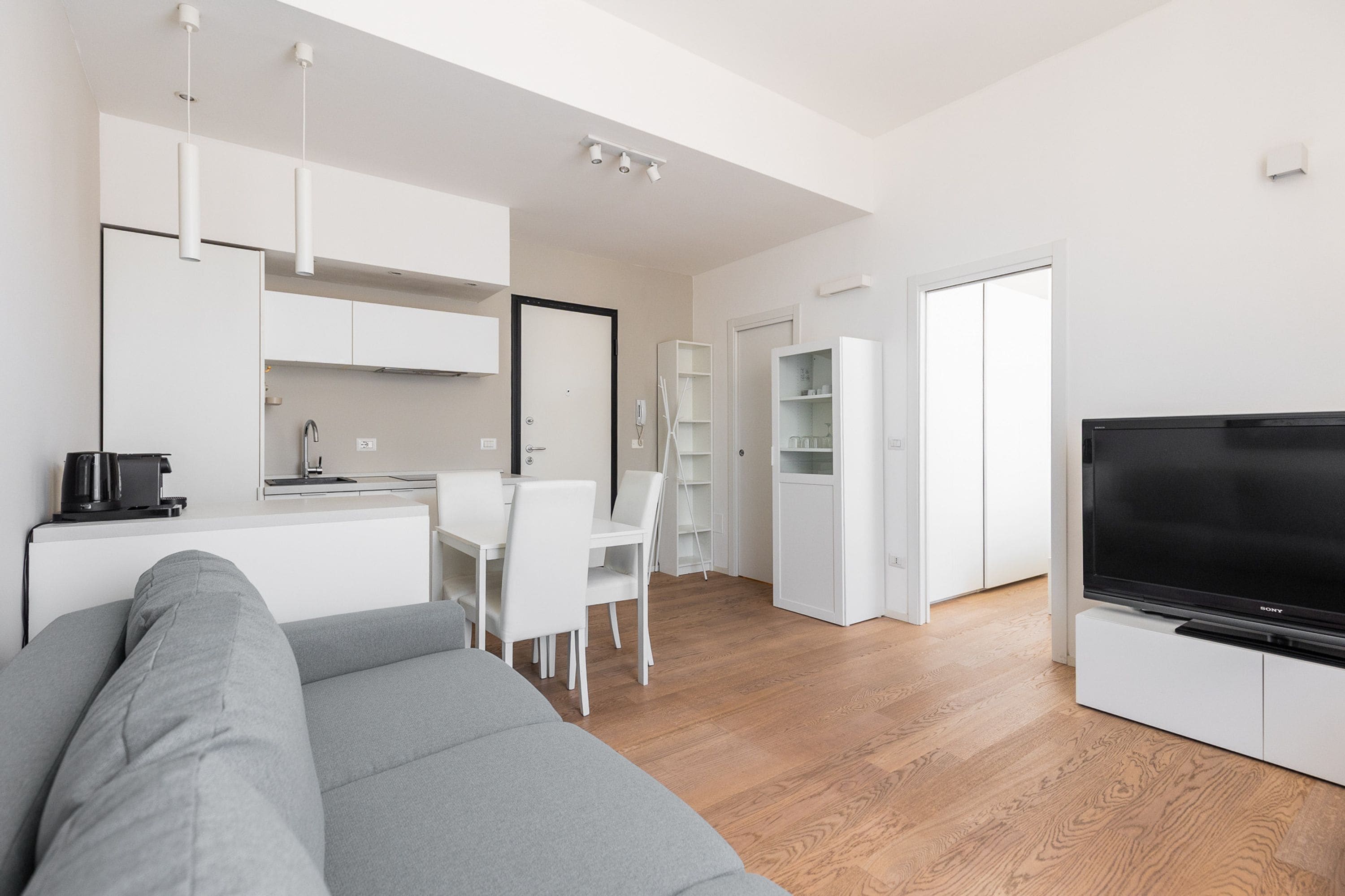 A modern living space with a grey sofa, a dining area with white chairs, a kitchen with white cabinets, and a black TV on a stand.