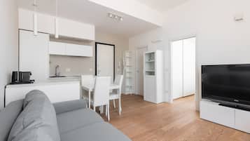 Een moderne woonruimte met een grijze bank, een eethoek met witte stoelen, een keuken met witte kasten en een zwarte televisie op een dressoir.