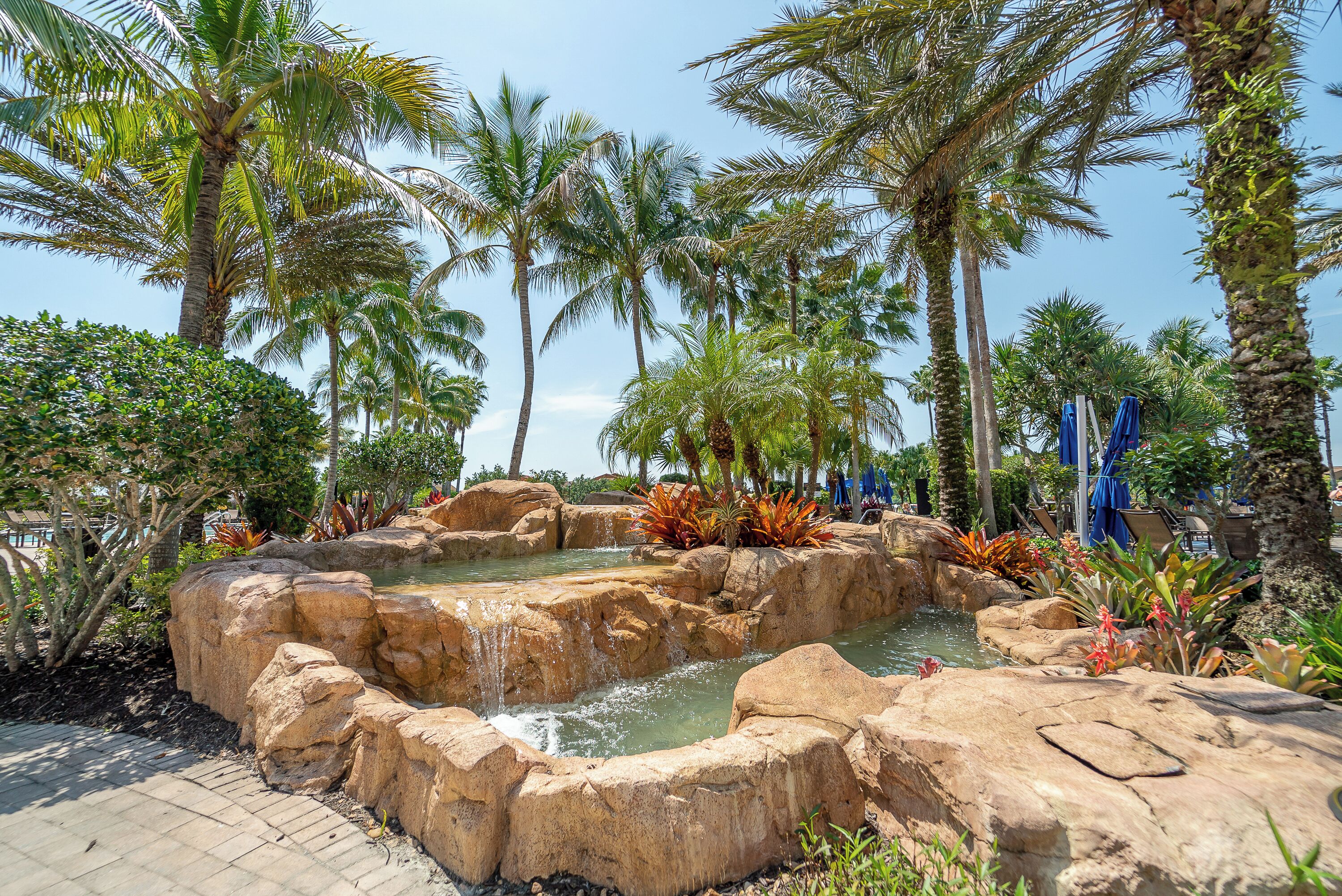 Een tropisch resort met een vijver met stenen, palmbomen en een verhard pad.