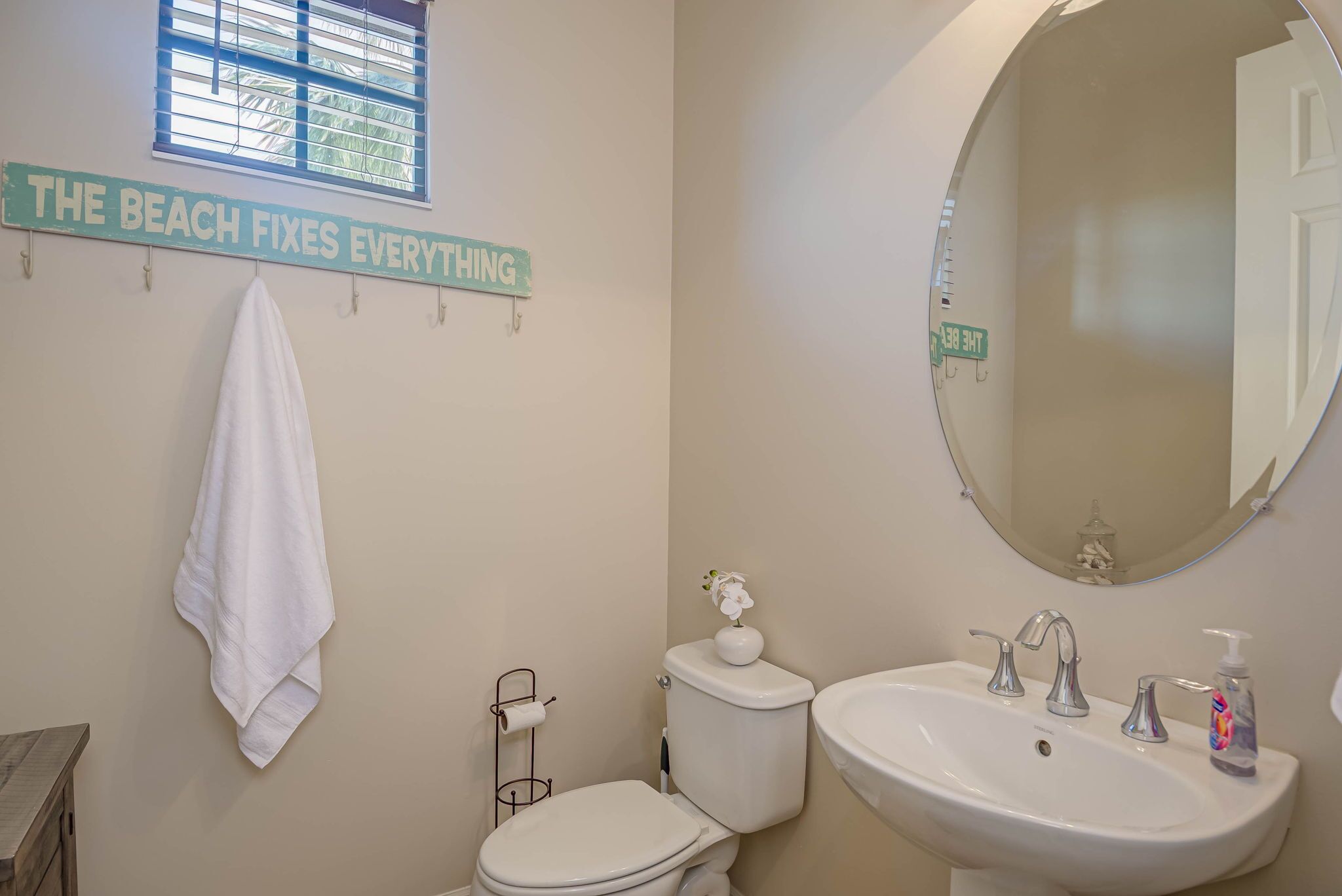 A bathroom with a toilet, sink, and a towel hanging on the wall. There is a window with blinds and a sign that reads 'THE BEACH FIXES EVERYTHING'.