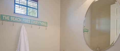 A bathroom with a toilet, sink, and a towel hanging on the wall. There is a window with blinds and a sign that reads 'THE BEACH FIXES EVERYTHING'.
